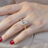 On hand shot of an emerald cut diamond bezel engagement ring. The bezel has a tulip engraving on the band. The hand has red nail polish and is resting on an arm.