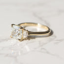 A yellow gold ring featuring a cushion-cut diamond center stone and a cadillac diamond side stone, resting on a white marble surface.
