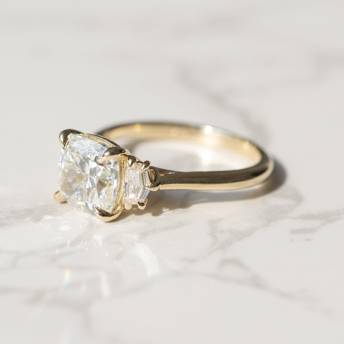 A yellow gold ring featuring a cushion-cut diamond center stone and a cadillac diamond side stone, resting on a white marble surface.