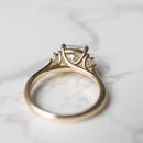 A gold ring with a clear cushion cut diamond in a three-stone setting, resting on a white marble surface.