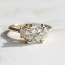 A yellow gold three-stone ring featuring a large cushion-cut center diamond flanked by two cadillac side diamonds, resting on a light marble surface.