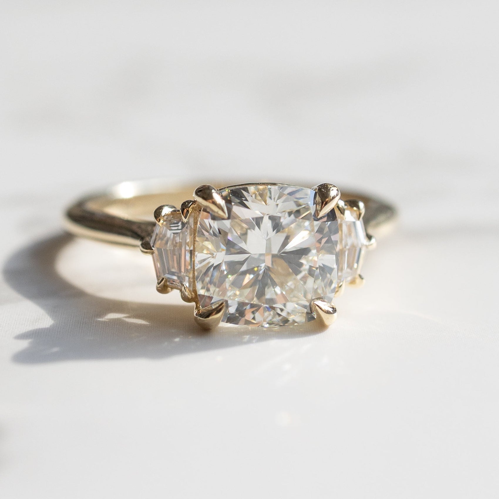 A yellow gold three-stone ring featuring a large cushion-cut center diamond flanked by two cadillac side diamonds, resting on a light marble surface.