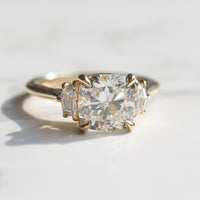 A yellow gold three-stone ring featuring a large cushion-cut center diamond flanked by two cadillac side diamonds, resting on a light marble surface.