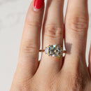 A close-up of a hand wearing a gold ring with a cushion-cut clear center diamond and smaller cadillac side stones, photographed against a light marble-like background.