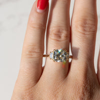 A close-up of a hand wearing a gold ring with a cushion-cut clear center diamond and smaller cadillac side stones, photographed against a light marble-like background.