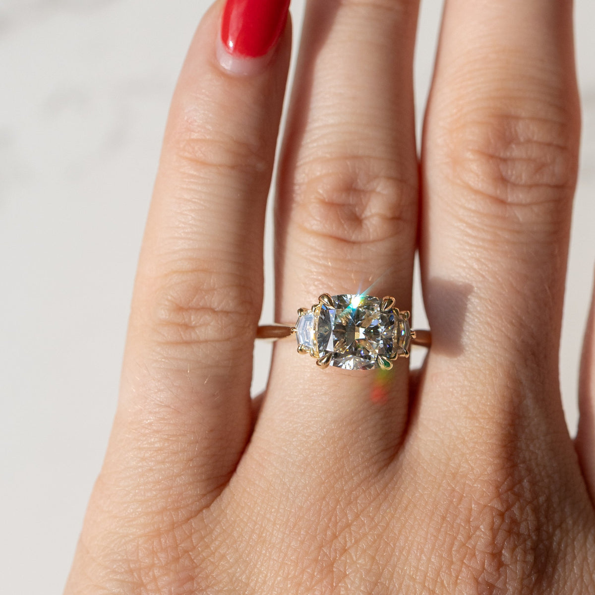 A close-up of a hand wearing a gold ring with a cushion-cut clear center diamond and smaller cadillac side stones, photographed against a light marble-like background.