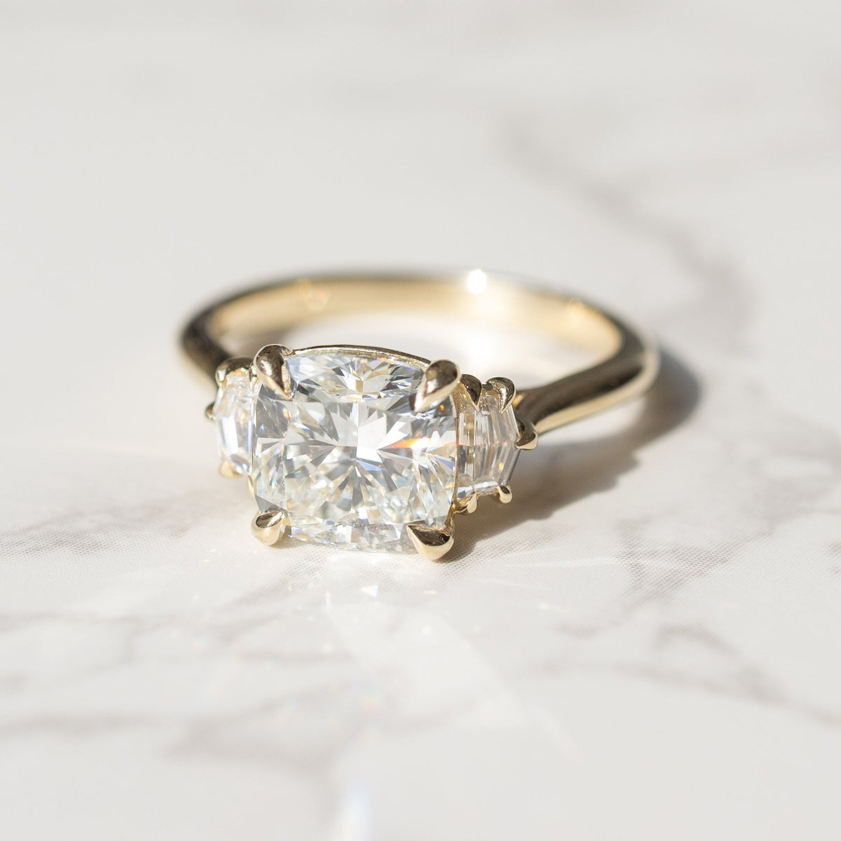 A yellow gold ring with a large cushion-cut diamond center stone and two smaller cadillac diamonds on either side, resting on a white marble surface.