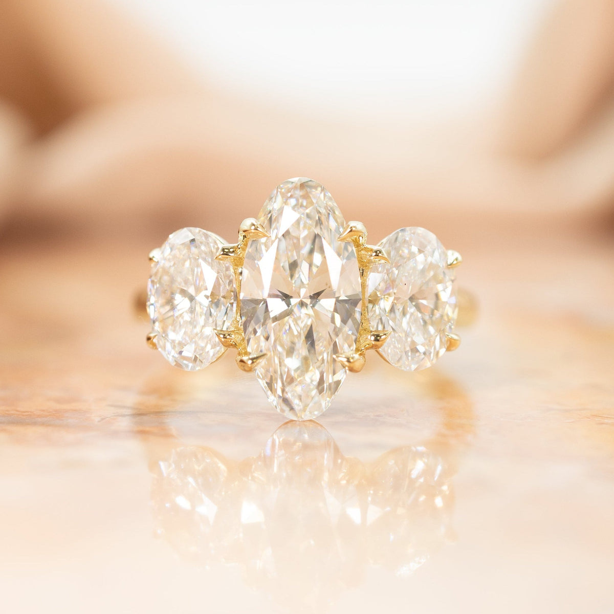 a large yellow gold three stone engagement ring with elongated ovals sitting on a blurred pink marble backdrop with fabric.