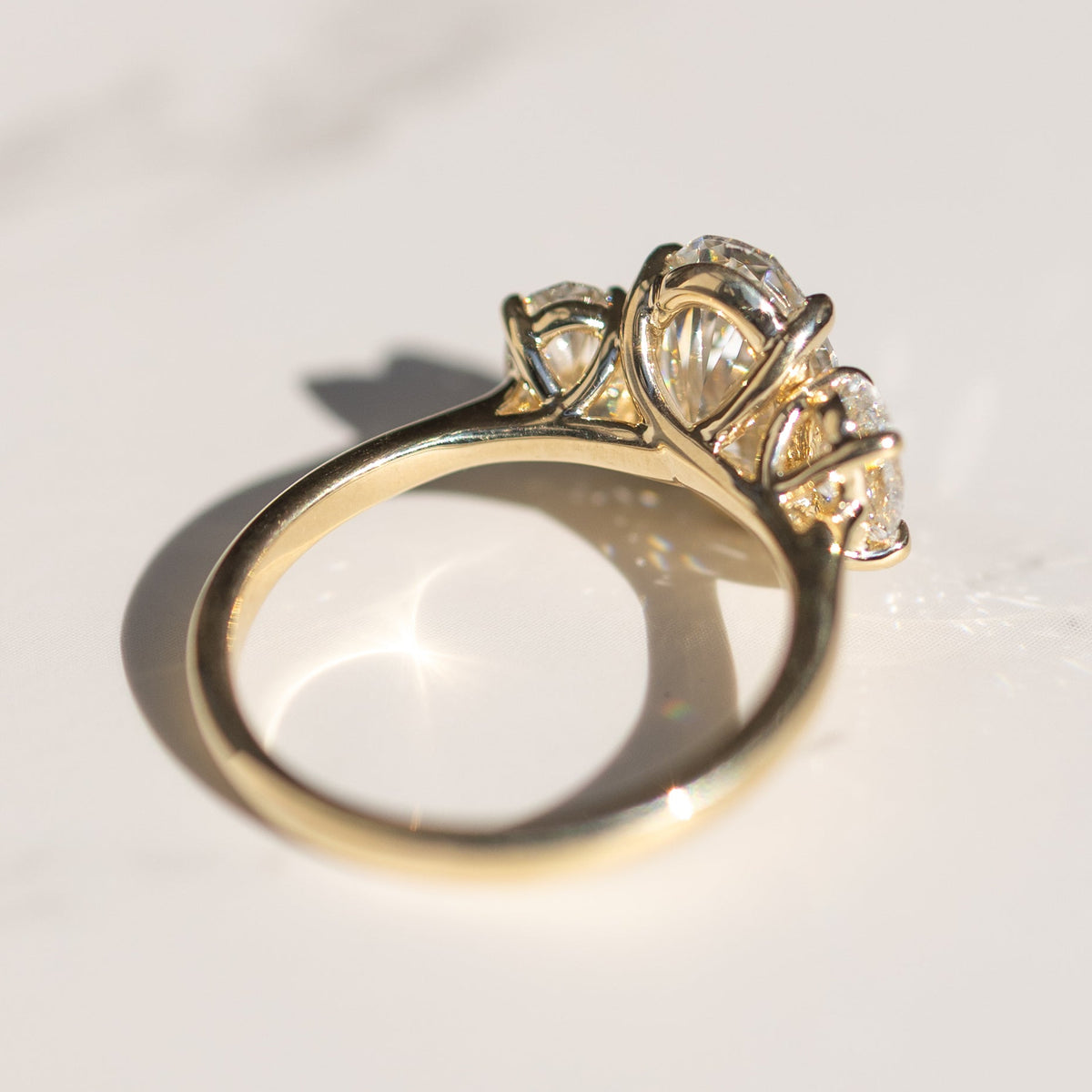 The bottom side of a yellow gold three stone engagement ring. The ring is sitting on a white marble backdrop with shadows.