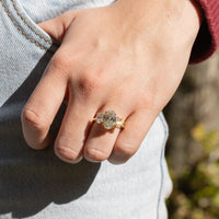 A woman's hand against blue jeans. On her left ring finger is a large oval triliogy engagement ring set in yellow gold. In the background is blurry green and brown foliage.