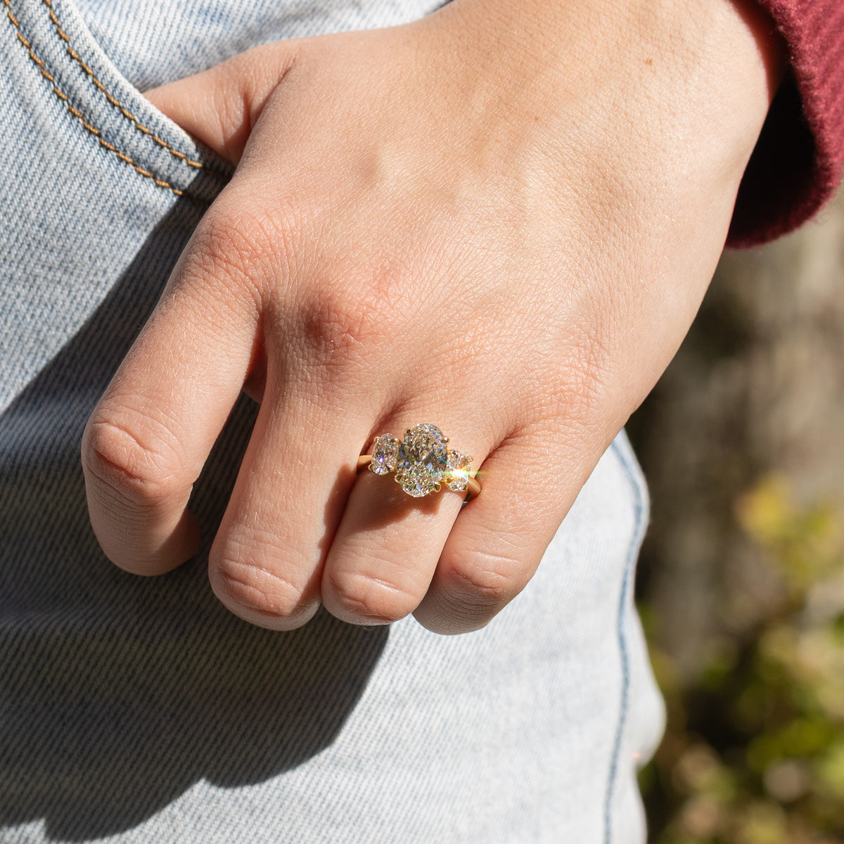 A woman's hand against blue jeans. On her left ring finger is a large oval triliogy engagement ring set in yellow gold. In the background is blurry green and brown foliage.