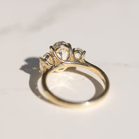 The back profile of a yellow gold three-stone oval engagement ring. The ring is sitting on a white marble backdrop.