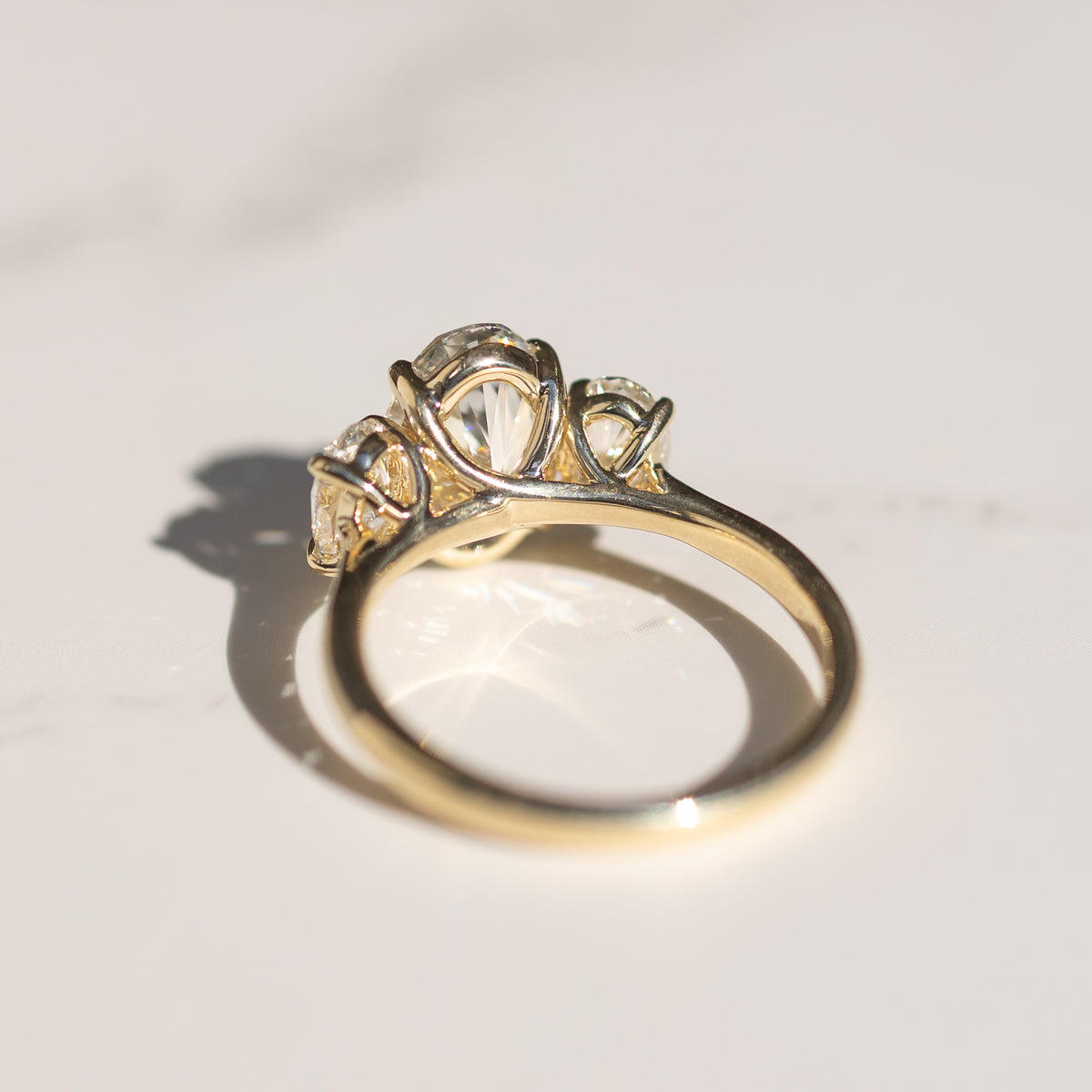 The back profile of a yellow gold three-stone oval engagement ring. The ring is sitting on a white marble backdrop.