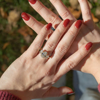 Two hands with red nail polish being held above blurry green and brown foliage. On the left hand's ring finger is a large three-stone oval engagement ring.