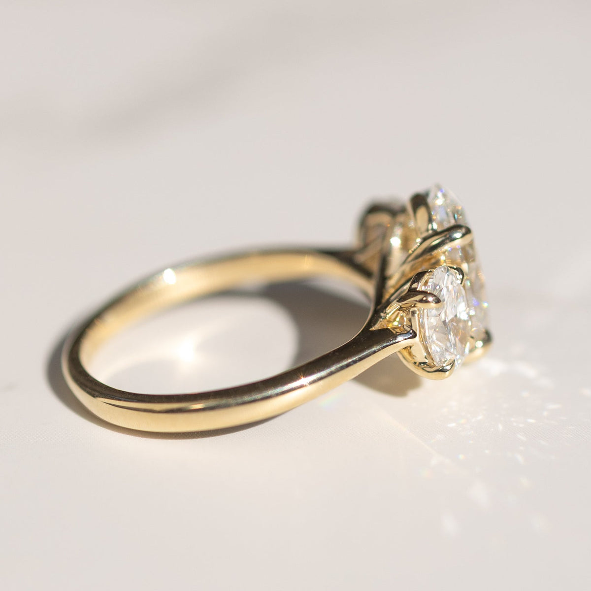 Side profile view of a three-stone yellow gold oval engagement ring. The ring is sitting on a white backdrop.