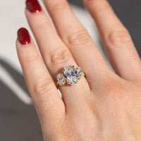 On-hand shot of a three-stone oval engagement ring set in yellow gold. The hand is being held in direct sunlight with a white backdrop behind it.