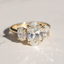 A three stone oval trilogy engagement ring sitting on a white marble backdrop in direct sunlight.