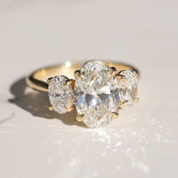 A three stone oval trilogy engagement ring sitting on a white marble backdrop in direct sunlight.