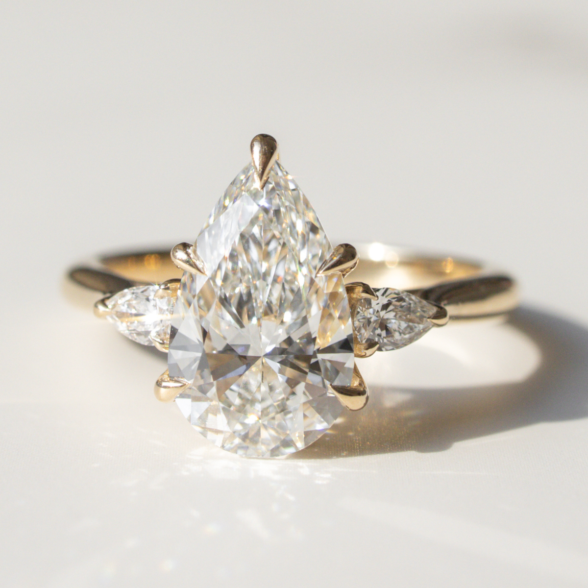 A pear cut three-stone engagement ring sitting on a white marble backdrop in sunlight.