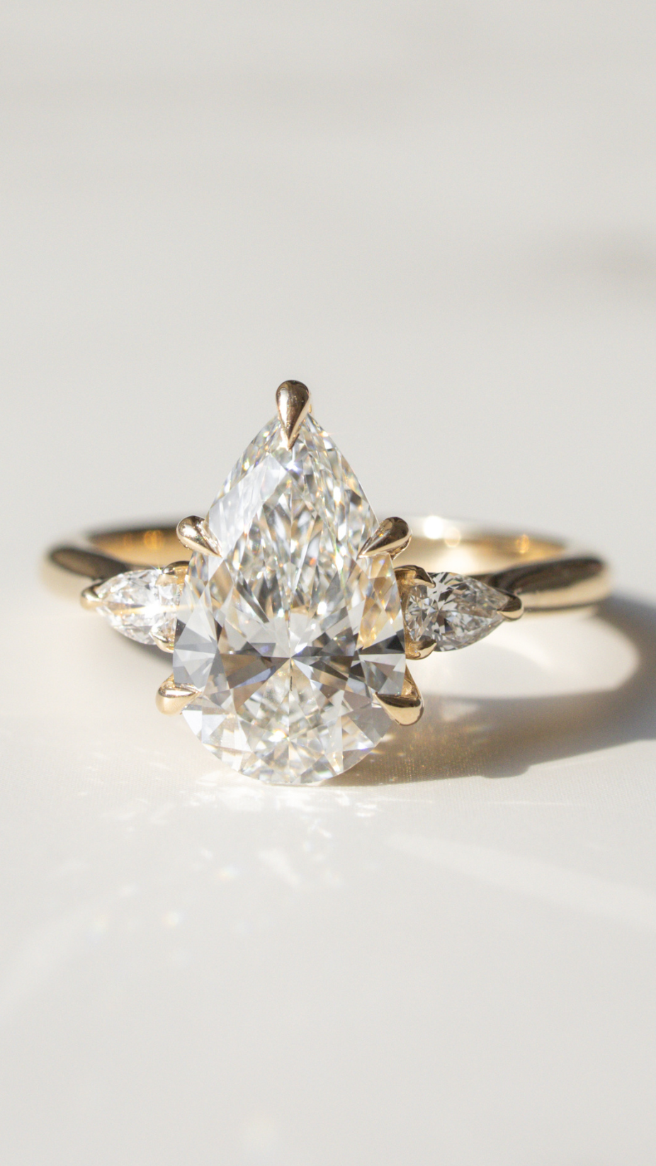 A large pear cut three-stone engagement ring set in yellow gold sitting on a white marble backdrop with shadows.