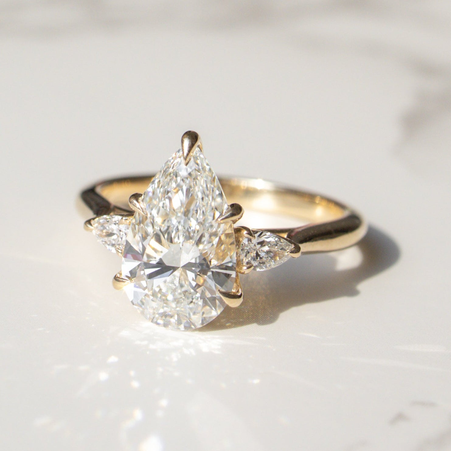 A three-stone gold ring featuring a pear-cut diamond center stone and smaller pear diamonds on the sides, resting on a white marble surface.