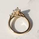 A yellow gold engagement ring with a pear diamond in a three-stone prong setting, photographed on a white marble surface with a soft shadow.