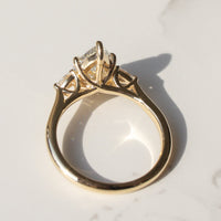 A yellow gold engagement ring with a pear diamond in a three-stone prong setting, photographed on a white marble surface with a soft shadow.