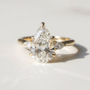 A gold ring featuring a pear-cut center diamond surrounded by pear-cut side diamonds, photographed on a light marble surface with a soft shadow.