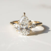 A gold ring featuring a pear-cut center diamond surrounded by pear-cut side diamonds, photographed on a light marble surface with a soft shadow.