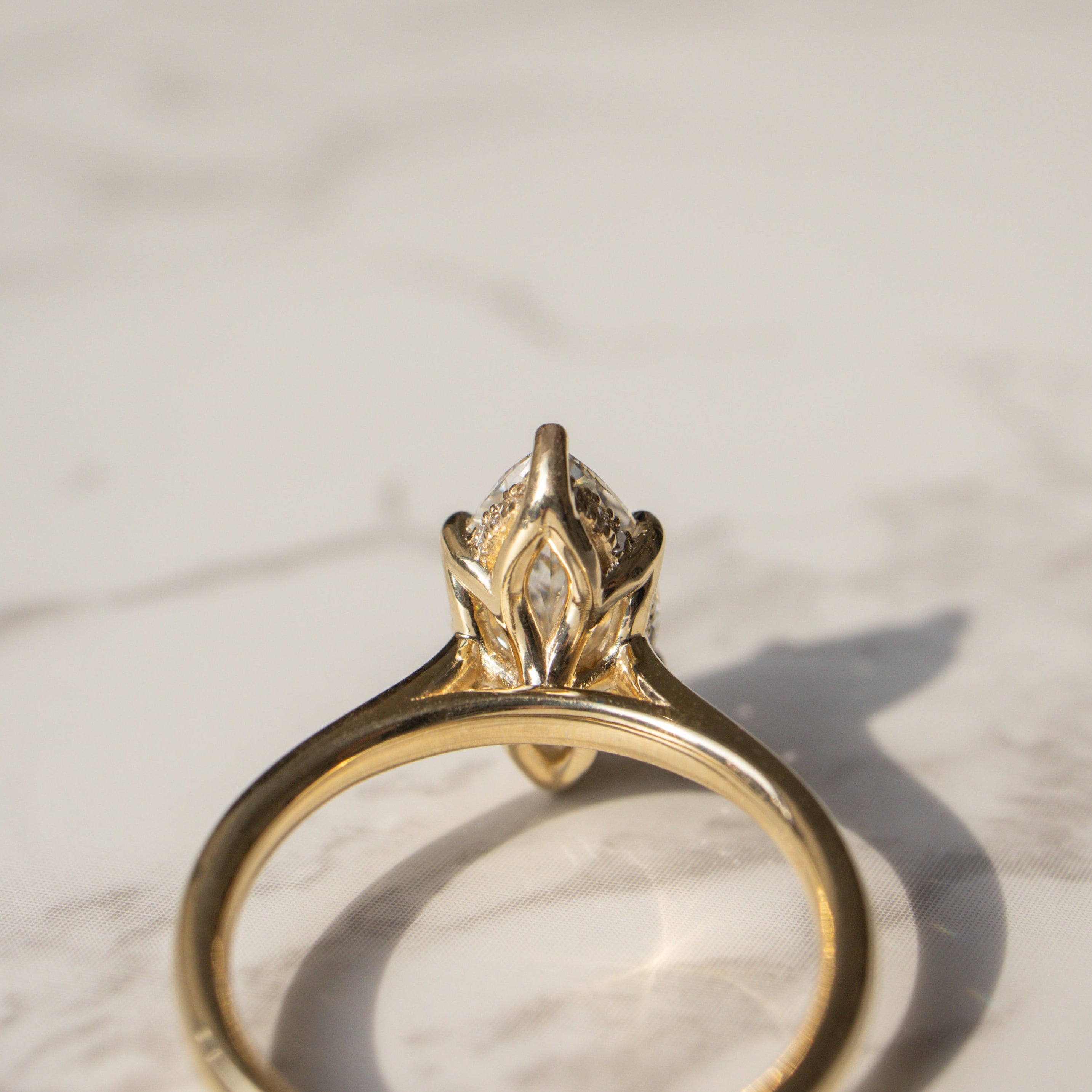 A gold ring with a marquise-cut diamond in a tulip hidden halo setting, resting on a light marble surface with a soft shadow.