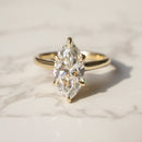 A yellow gold ring with a marquise-cut diamond solitaire setting, resting on a light marble surface.