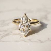 A yellow gold ring with a marquise-cut diamond solitaire setting, resting on a light marble surface.