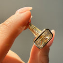 A close-up of a hand holding a gold ring with a large emerald-cut diamond. The ring features an intricate floral pattern on the band and a bezel-set diamond setting.