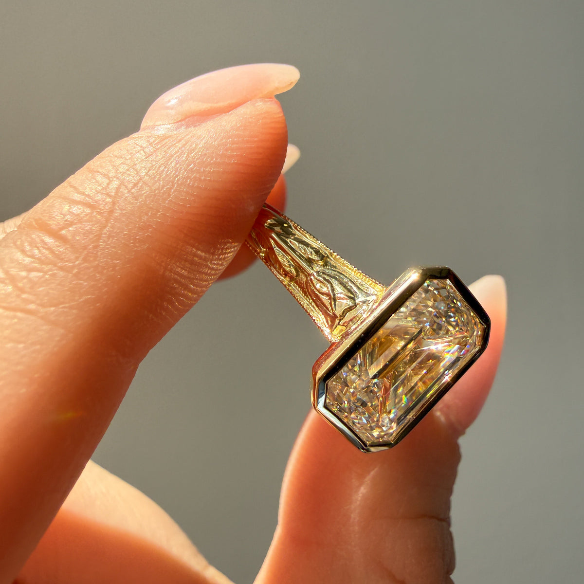 A close-up of a hand holding a gold ring with a large emerald-cut diamond. The ring features an intricate floral pattern on the band and a bezel-set diamond setting.