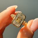 A close-up of a hand holding a gold ring with a large emerald-cut diamond. The ring features a yellow gold band with intricate engravings and a prominent emerald-cut center stone.