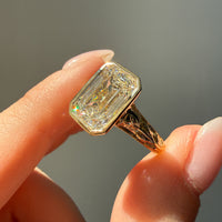 A close-up of a hand holding a gold ring with an emerald-cut diamond. The ring features an engraved band and is photographed against a soft, out-of-focus gray background.