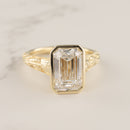A yellow gold ring featuring a large rectangular emerald-cut clear diamond in a bezel setting, with an ornate openwork gold band, shown on a light marble surface.