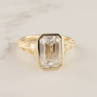 A yellow gold ring featuring a large rectangular emerald-cut clear diamond in a bezel setting, with an ornate openwork gold band, shown on a light marble surface.