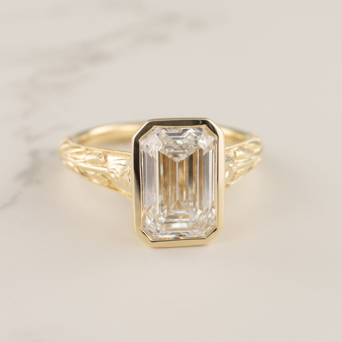 A yellow gold ring featuring a large rectangular emerald-cut clear diamond in a bezel setting, with an ornate openwork gold band, shown on a light marble surface.