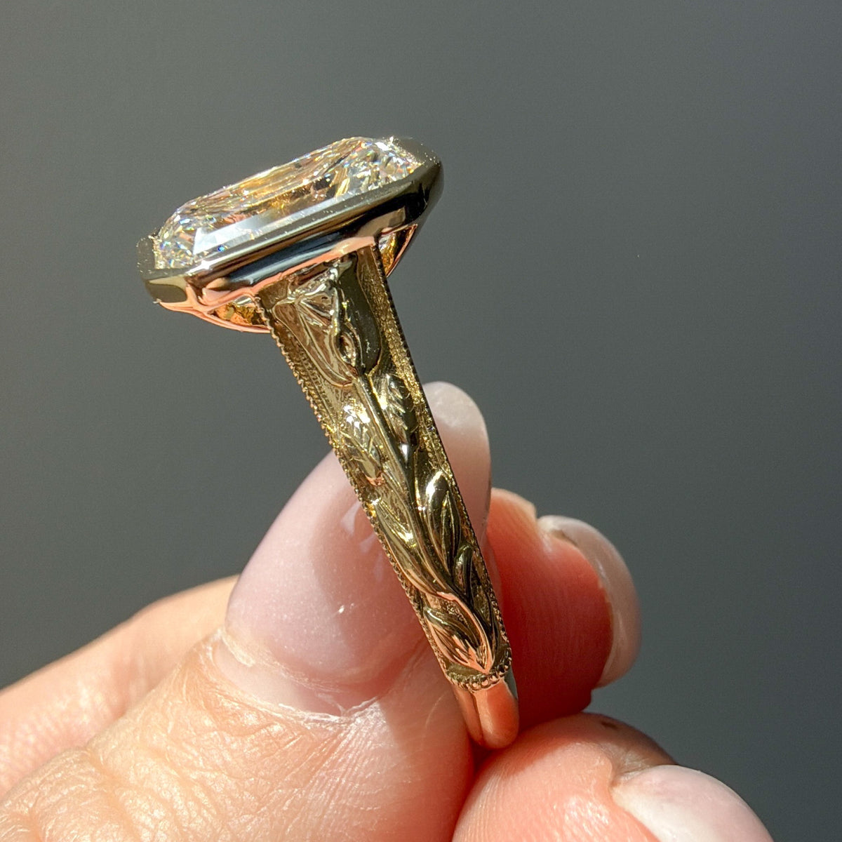 An antique-style gold ring with a large emerald diamond and an intricately engraved shank, held between fingers against a dark gray background.