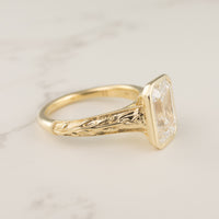 A bezel-set engagement ring with tulip floral filigree along the shank on a white marble backdrop