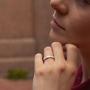A close-up lifestyle shot of a woman resting her chin on her hand, wearing a delicate yellow gold ring with small baguette diamonds on her ring finger. The background is a softly blurred brick wall.