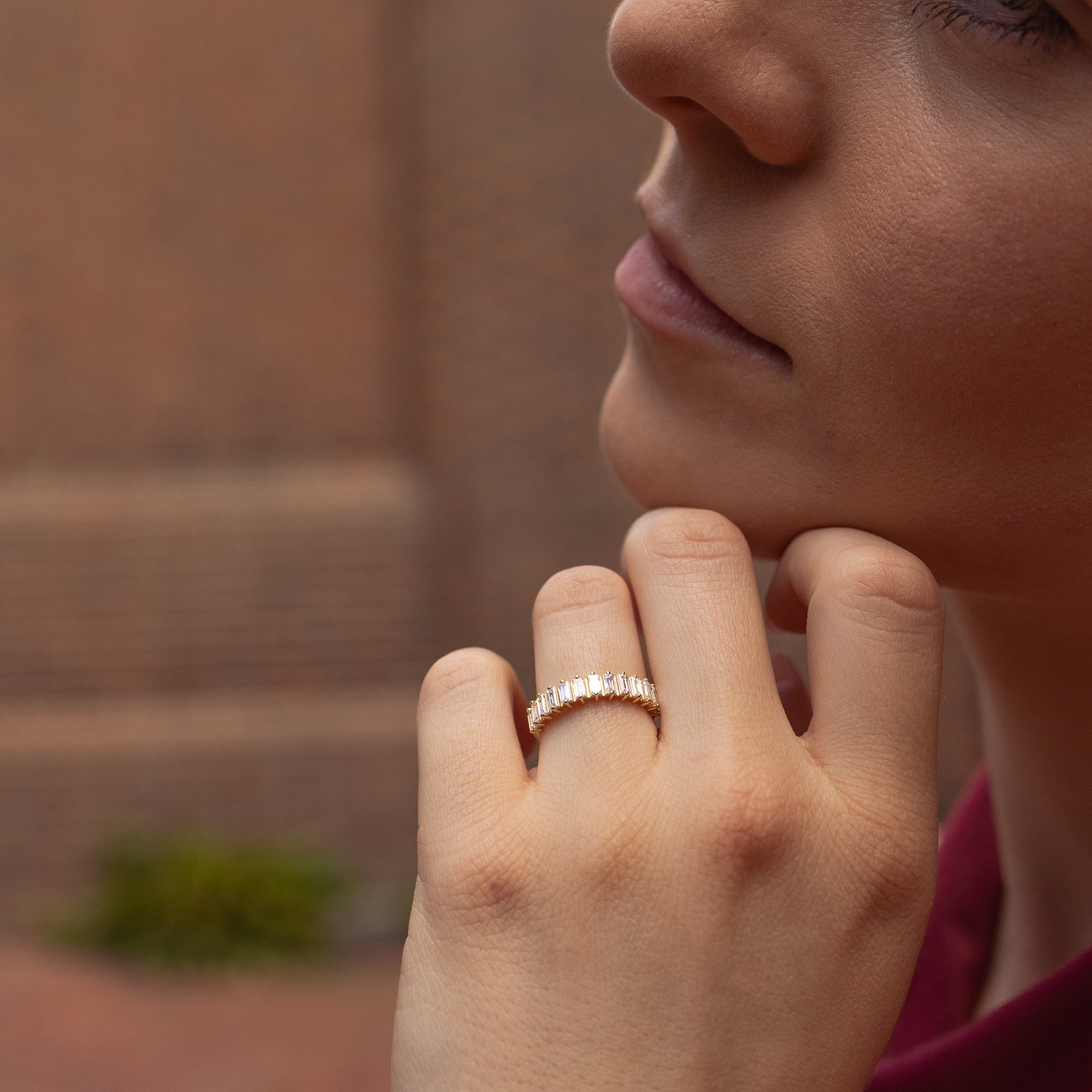 A close-up lifestyle shot of a woman resting her chin on her hand, wearing a delicate yellow gold ring with small baguette diamonds on her ring finger. The background is a softly blurred brick wall.