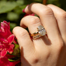 A close-up of a hand wearing a yellow gold ring with a large oval-cut diamond center stone and a wedding band with smaller baguette diamonds, set against a blurred background of green foliage and bright pink flowers.