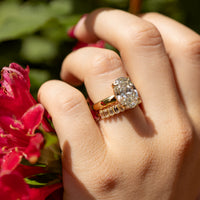 A close-up of a hand wearing a yellow gold ring with a large oval-cut diamond center stone and a wedding band with smaller baguette diamonds, set against a blurred background of green foliage and bright pink flowers.