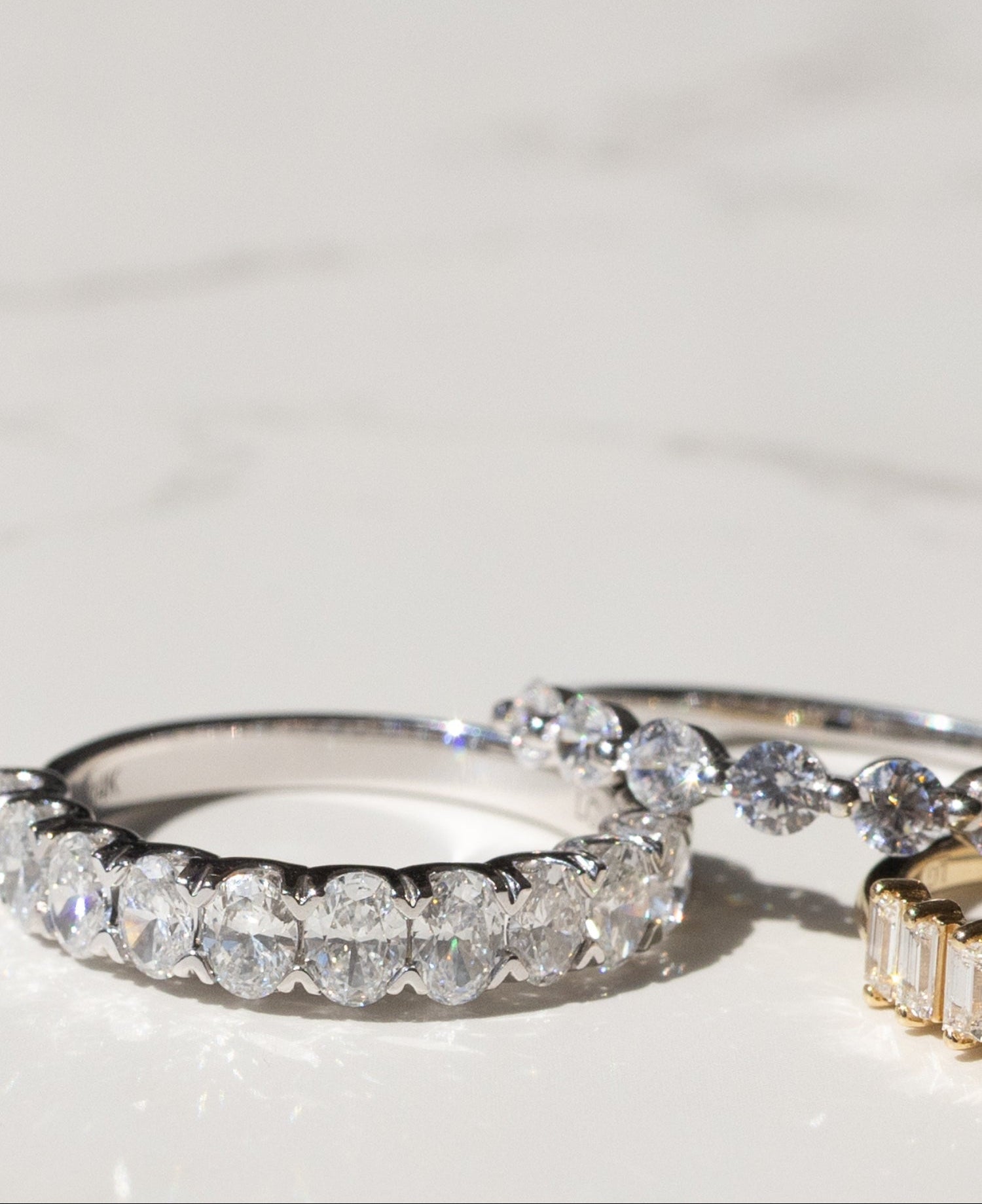 A close-up of a silver-toned ring set with oval brilliant-cut diamonds, photographed on a light marble-like surface. Part of a gold-toned ring is visible at the right edge.