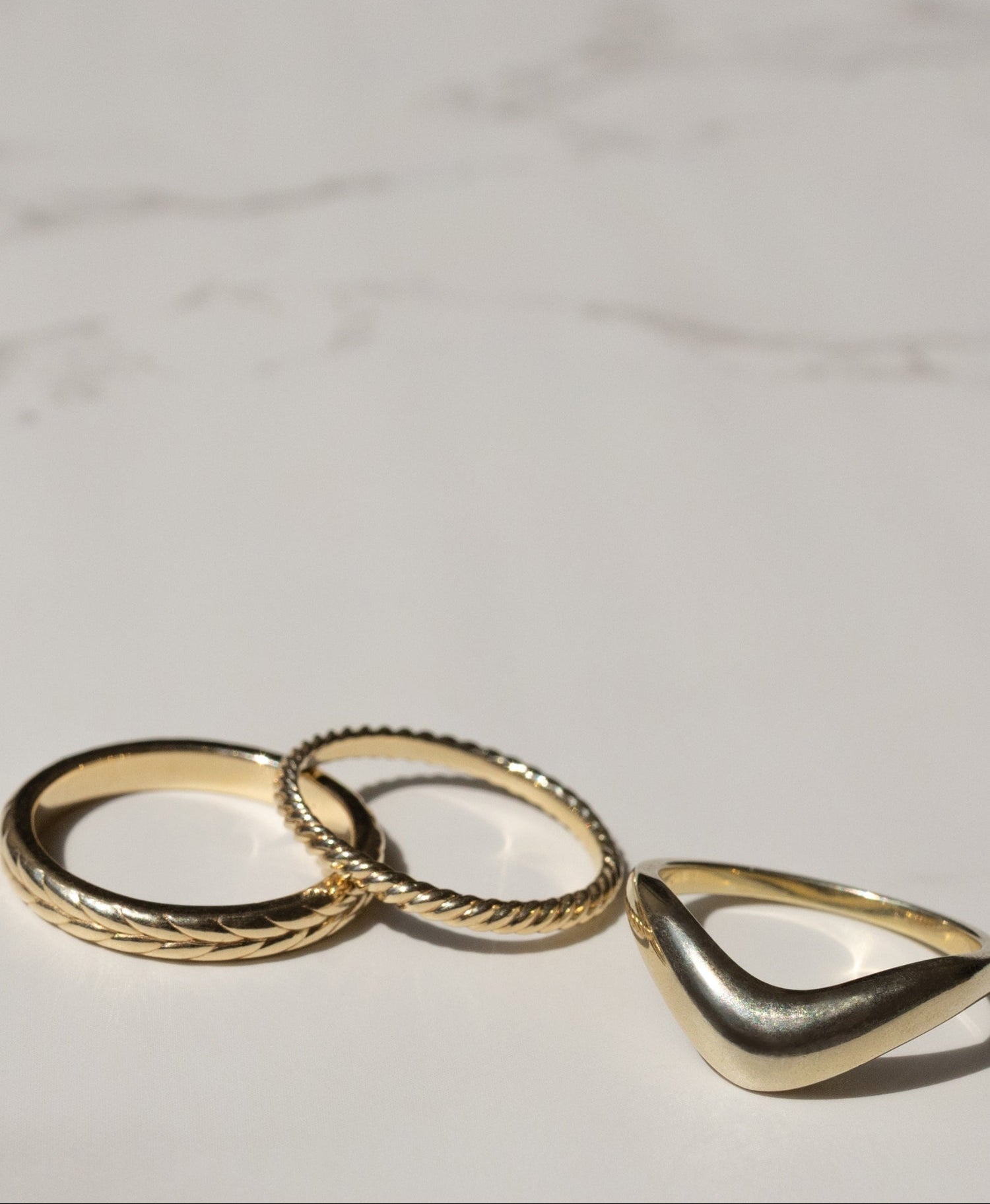 Three gold rings displayed on a light marble surface, including two overlapping textured rings and one triangular ring.
