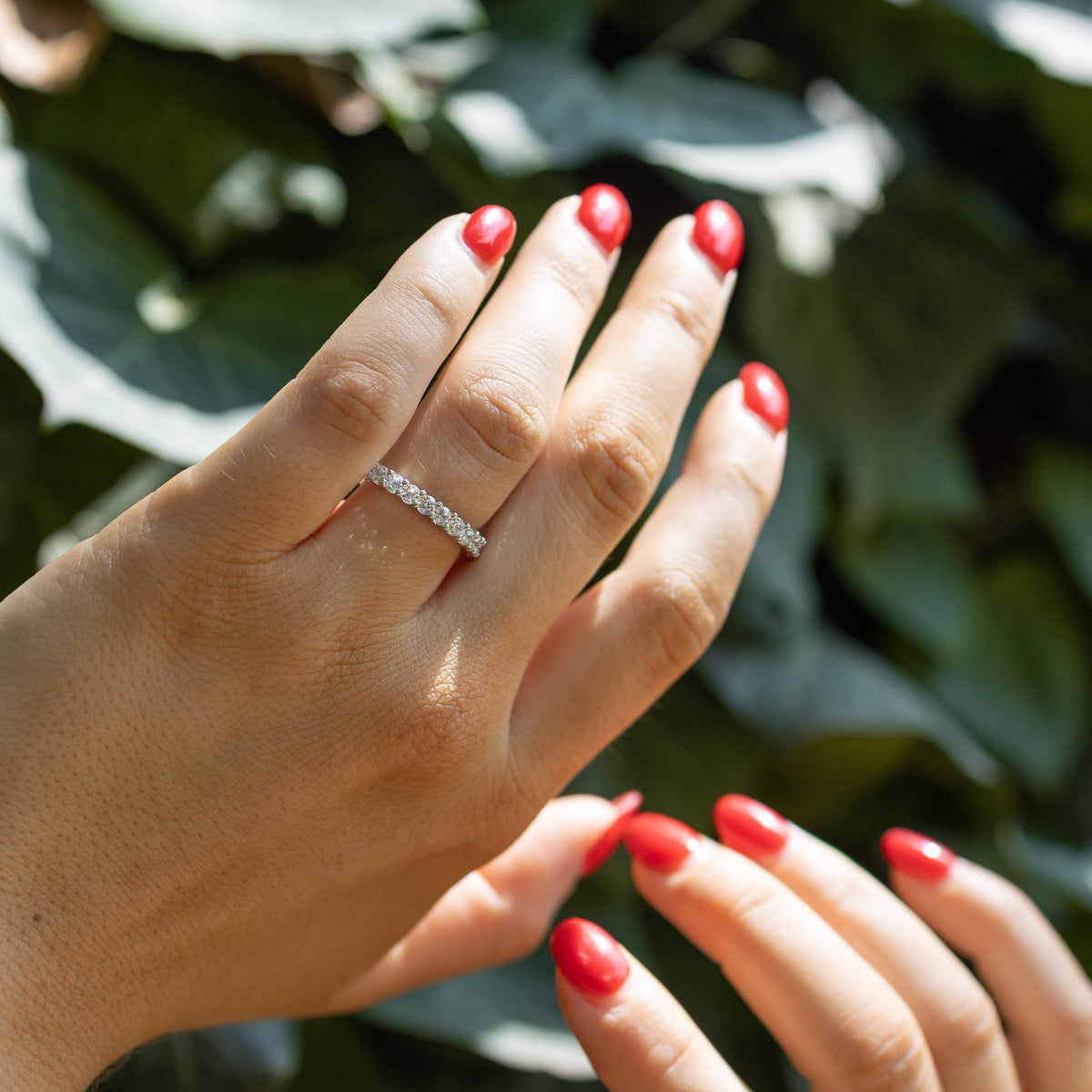 A close-up of two hands with bright red manicured nails, with one hand wearing a delicate white gold diamond ring. The background is composed of lush, out-of-focus green leaves.