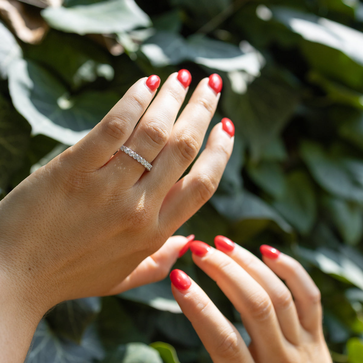 A close-up of two hands with bright red manicured nails, with one hand wearing a delicate white gold diamond ring. The background is composed of lush, out-of-focus green leaves.