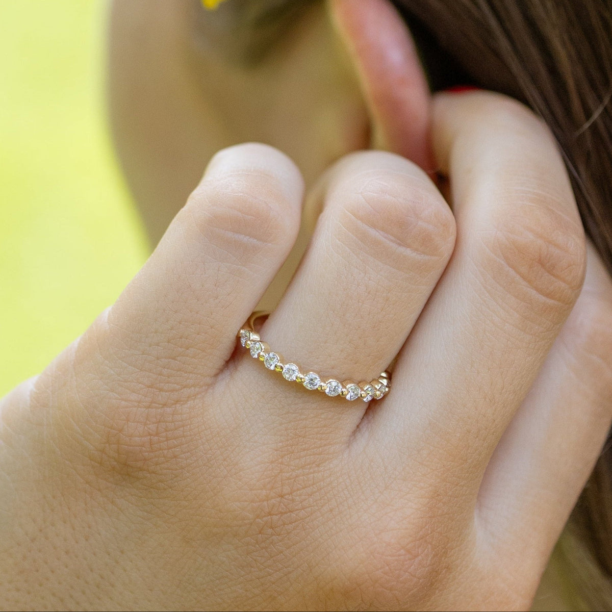 A close-up lifestyle shot of a woman's hand wearing a delicate gold ring with small clear stones, holding a small yellow flower behind her ear. The background is a soft-focus green outdoor lawn.